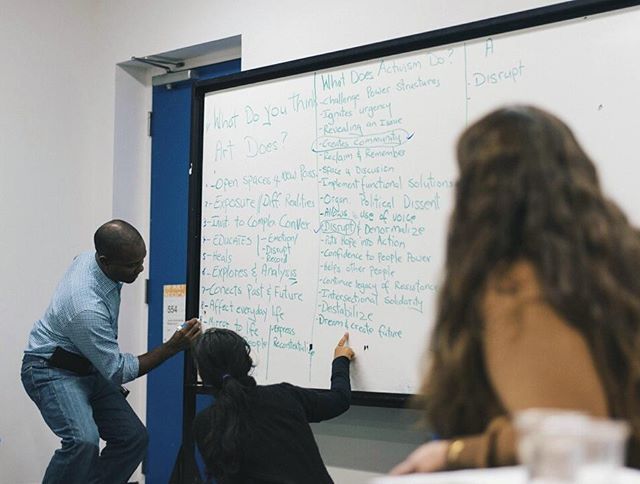 Two people writing on a whiteboard; someone on the right watching them, back turned to the camera.