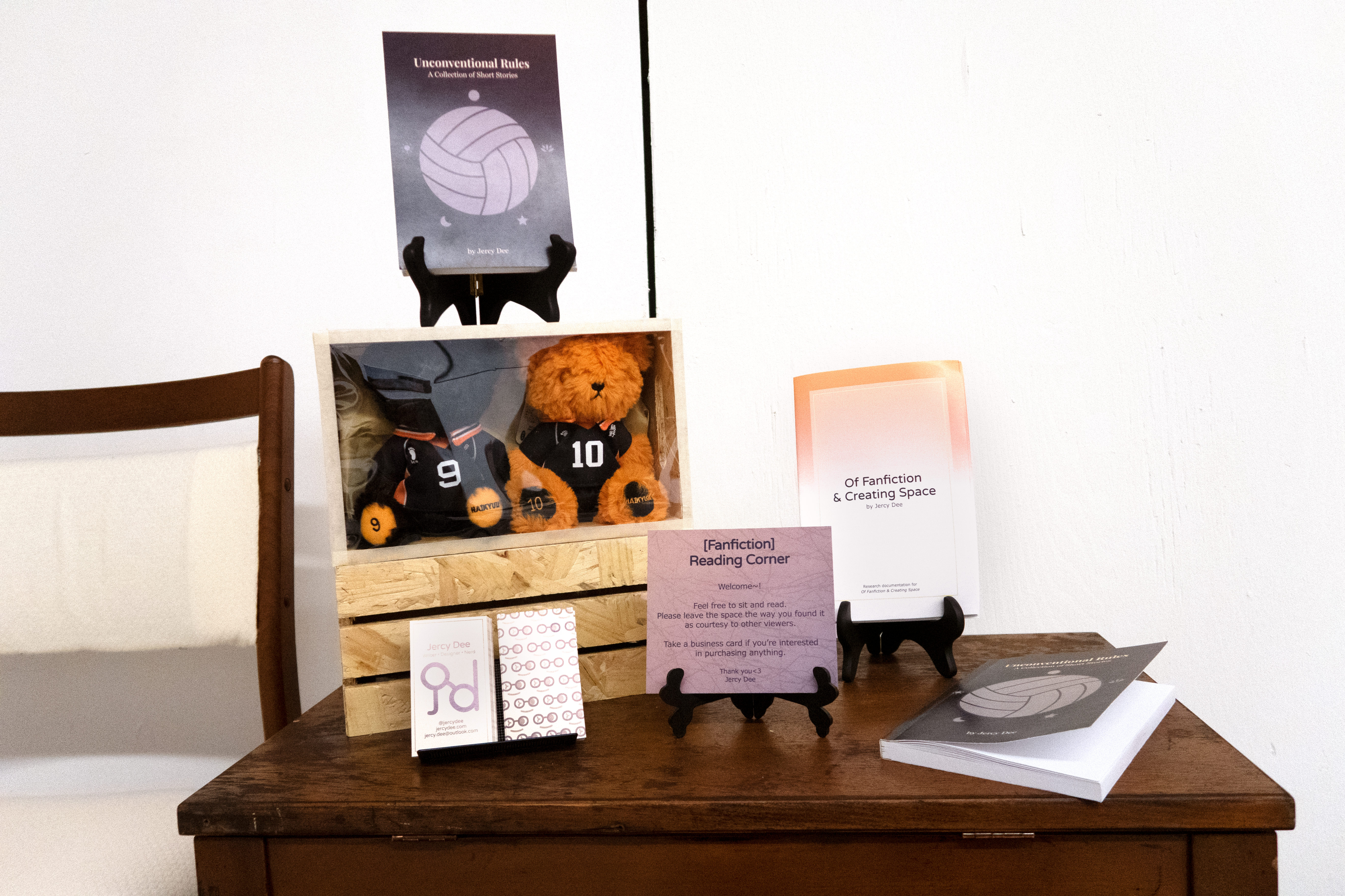 A close-up shot of the reading space, specifically of the contents on the table. From left to right: decorative boxes with one copy of Unconventional Rules standing on top, a printed copy of OFACS primary research documentation standing next to the boxes, and the other copy of Unconventional Rules lying next to it. Also on the table are business cards and a welcome sign for the viewers.